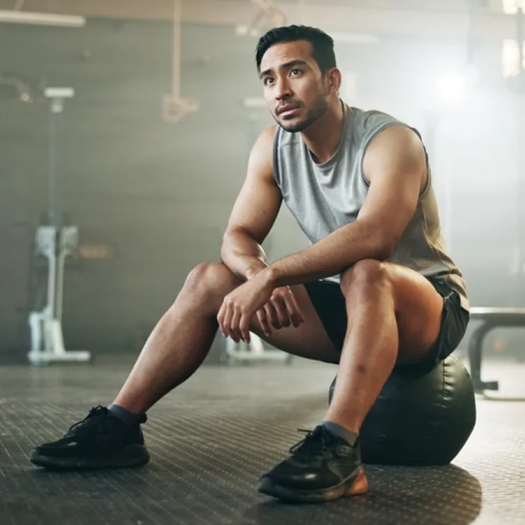 A man wearing a tank top and gym shorts, sits on a workout ball in a gym.